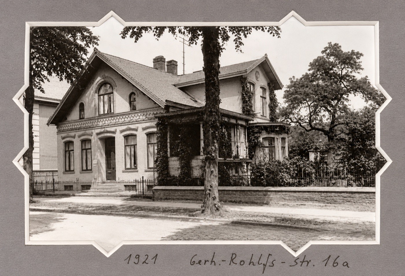 Schwarz-Weiß-Foto von 1921: ein zweistöckiges Haus mit Giebeln, Veranda und Vorgarten, davor ein Baum. Darunter handschriftlich: 1921, Gerh.-Rohlfs-Str. 16a.