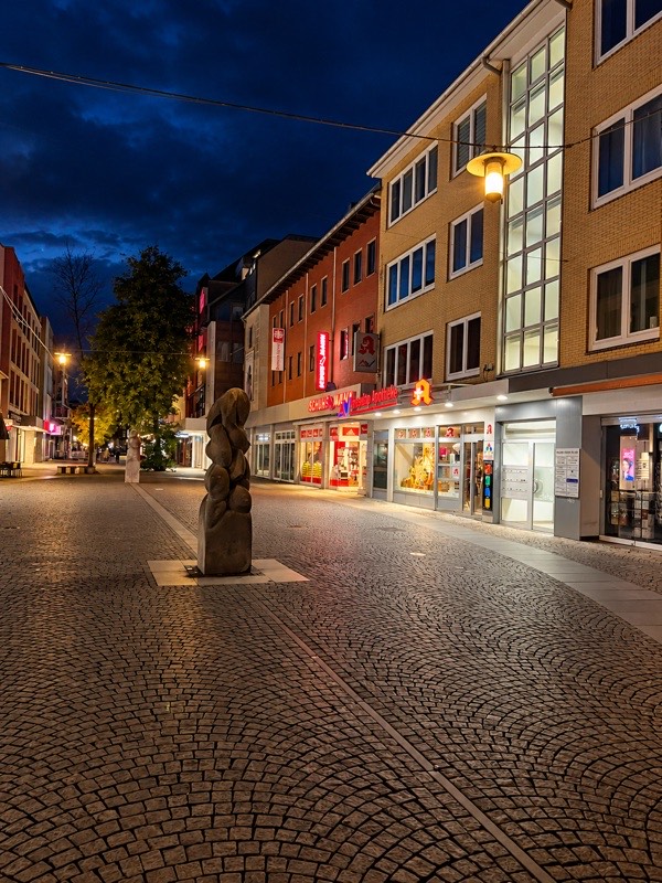 Die Gerhard-Rohlfs-Straße heute am Abend: Fußgängerzone mit Kopfsteinpflaster, im Erdgeschoss unseres Hauses die Apotheke.